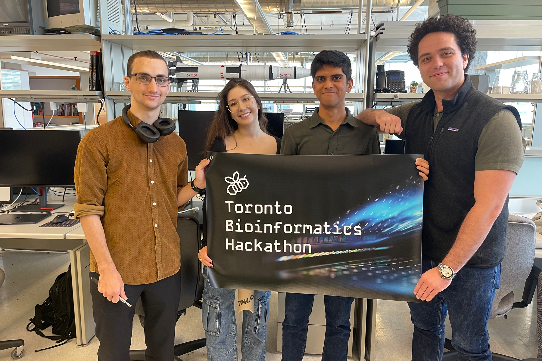 Four people standing in a computer lab holding a sign that says "Toronto Bioinformatic Hackathon"