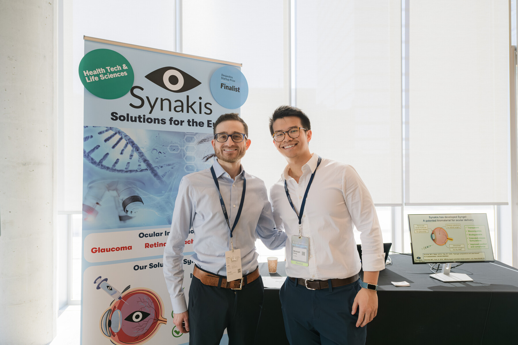 Two Synakis team members standing in front of a table and pull-up banner for their company