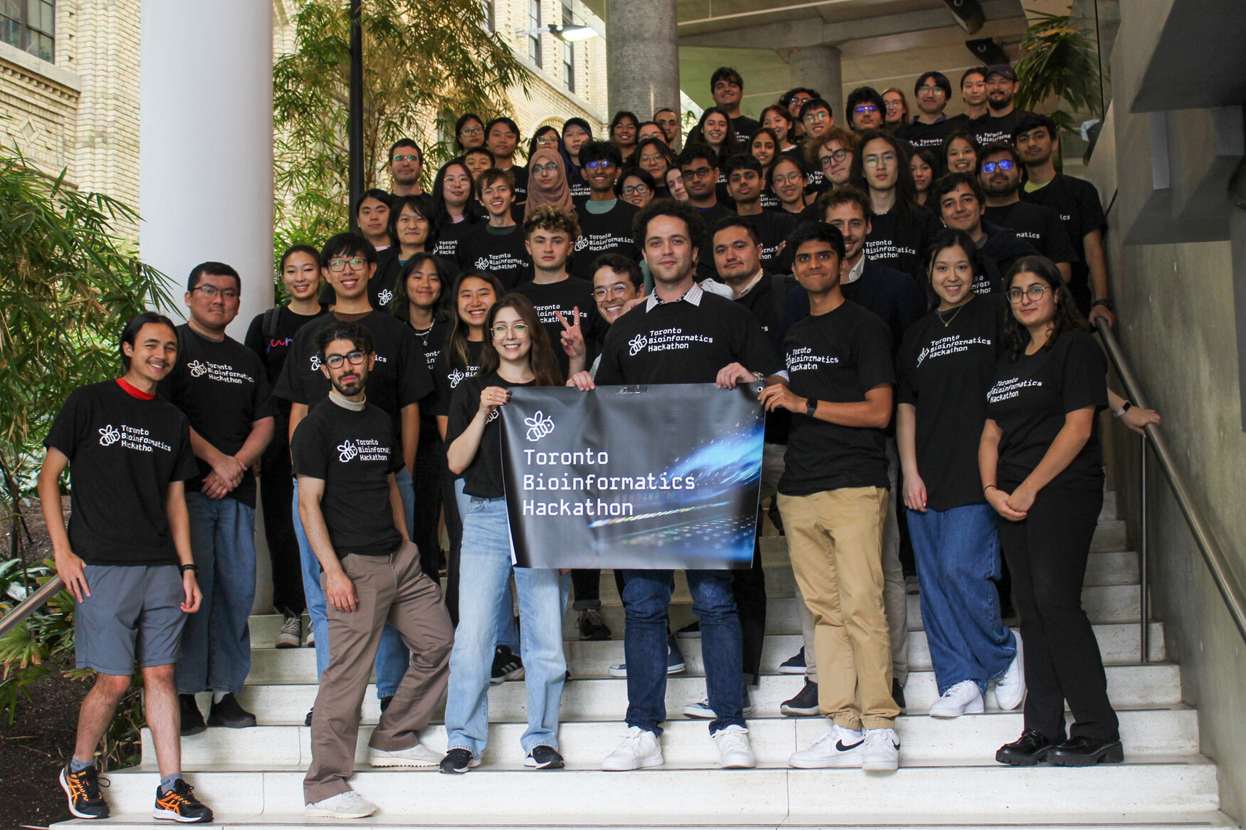 A group of around 50 people in black T-shirts branded with "Toronto Bioinformatics Hackathon" standing on a staircase